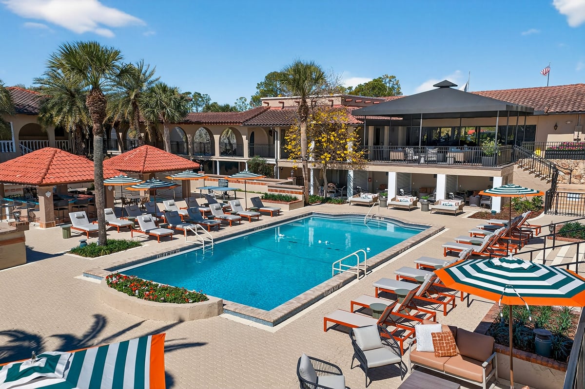 A luxurious resort pool with lounge chairs, colorful umbrellas, and palm trees, surrounded by Spanish-style architecture on a sunny day.