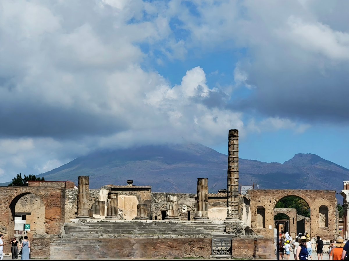 Ancient Roman ruins with towering columns and stone arches stand in the foreground, with Mount Vesuvius visible in the background under a cloudy blue sky.