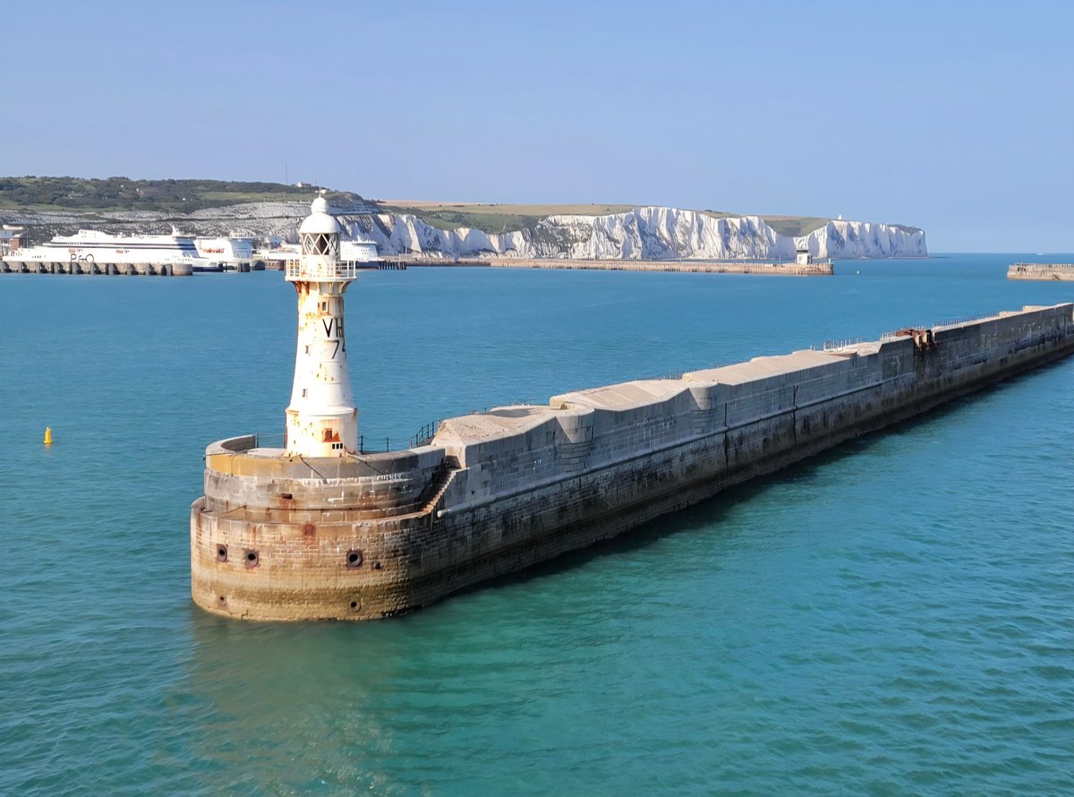A white lighthouse with a keeper's cottage stands at the end of a long stone breakwater extending into turquoise water, with white chalk cliffs visible across the bay.
