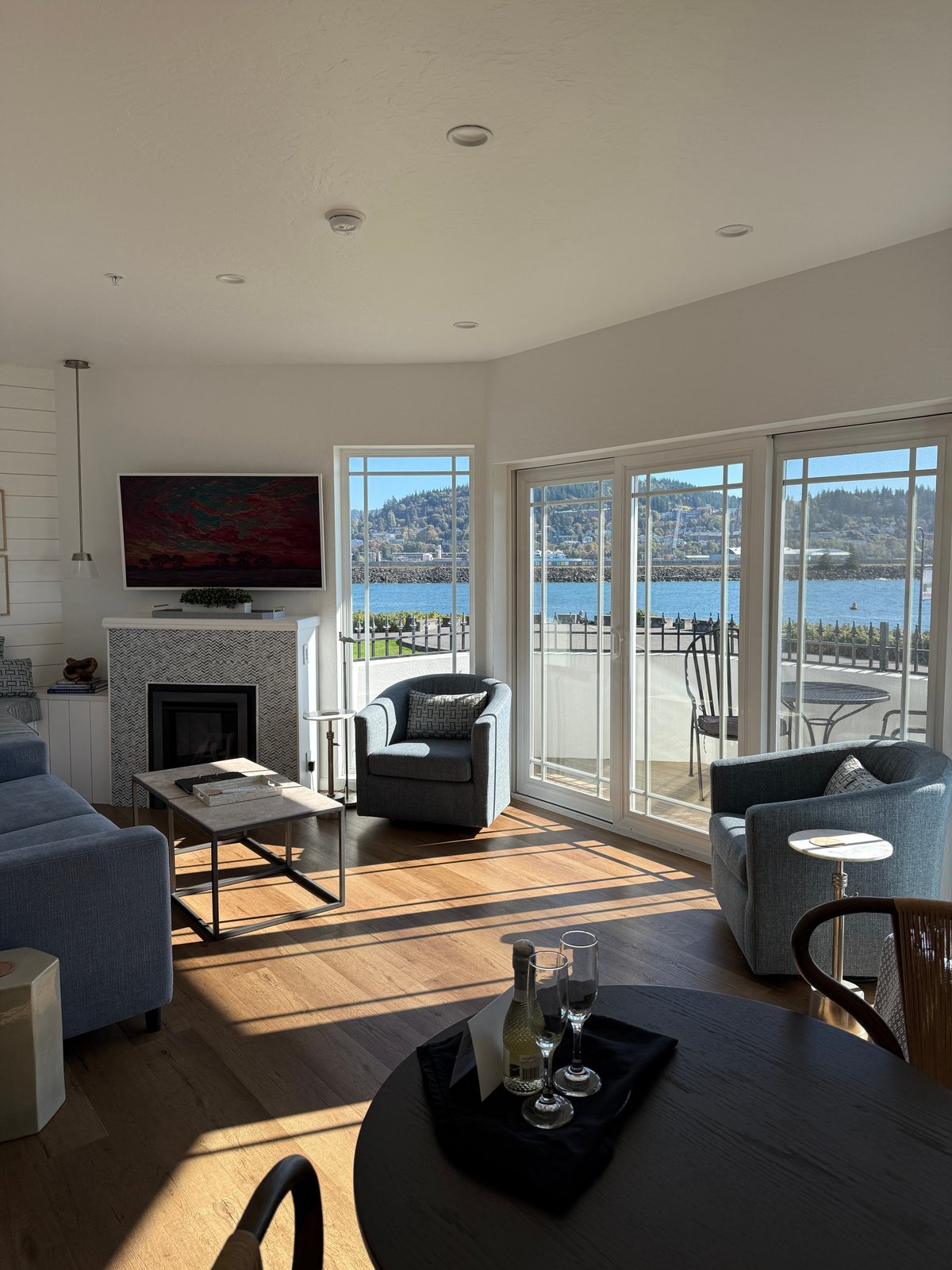 A bright, modern waterfront living room with floor-to-ceiling windows overlooking a lake surrounded by forested mountains.