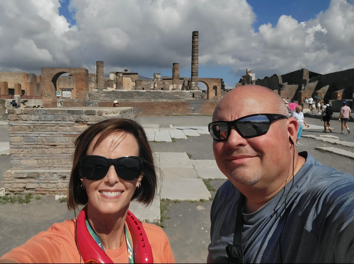 Two visitors pose for a selfie at the ancient Roman ruins of Pompeii, with excavated columns and structures visible behind them under a cloudy blue sky.