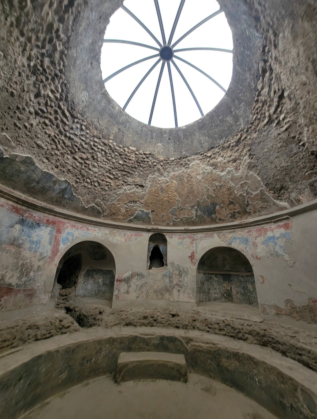 Interior view of a Roman bathhouse with circular skylight showing geometric spokes, arched alcoves, and deteriorated frescoed walls revealing brick beneath.