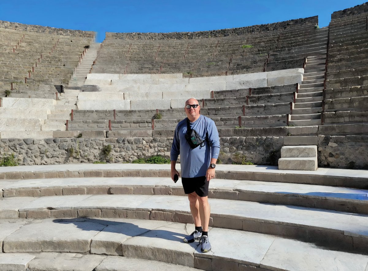 A jogger in casual athletic wear stands on ancient stone amphitheater steps beneath a clear blue sky.