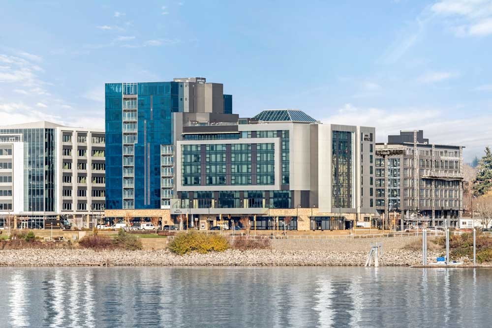 Modern riverfront development with mixed-use buildings featuring teal, white, and gray facades overlooking calm water on a clear day.