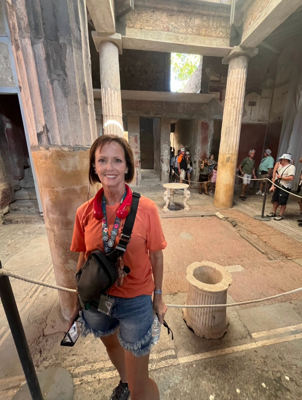 A woman in an orange shirt smiles at the camera inside an ancient Roman building with large fluted columns and a central courtyard.