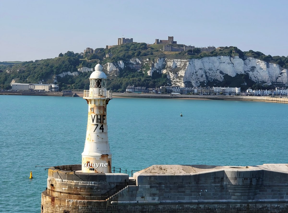 White lighthouse with VHF 74 marking stands on a stone breakwater in turquoise waters, with white chalk cliffs and a hilltop castle visible across the bay.