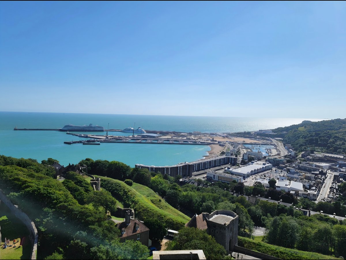 Aerial view of a coastal town with turquoise waters, cruise ships at dock, historic castle ruins on lush green hillside, and modern beachfront developments.