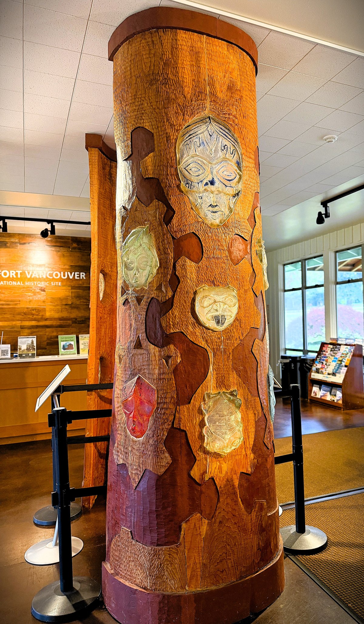 A tall wooden carved column featuring multiple artistic faces and figures in a museum or cultural center, with gallery signage visible in the background.