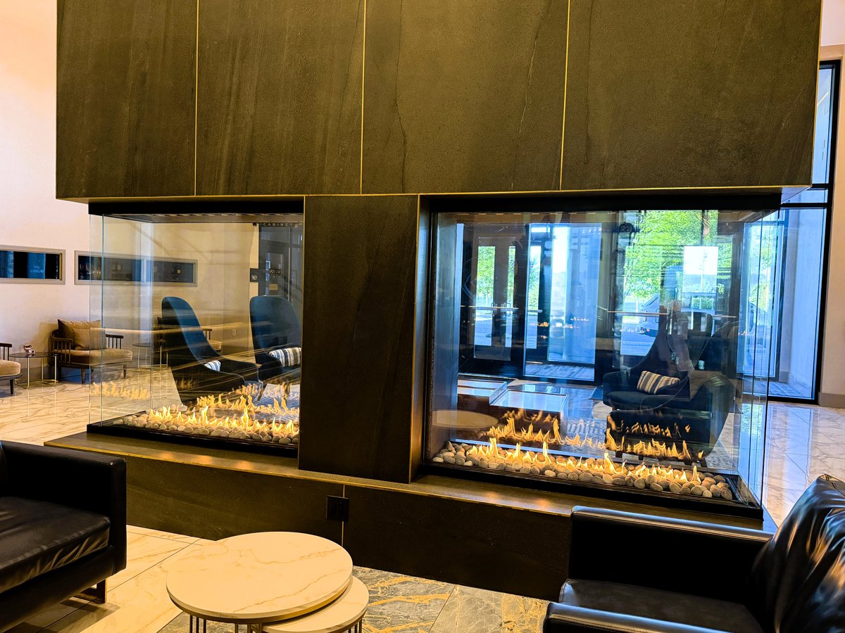 A modern luxury lobby with a two-sided gas fireplace, floor-to-ceiling windows overlooking green spaces, black leather seating, and contemporary furnishings.