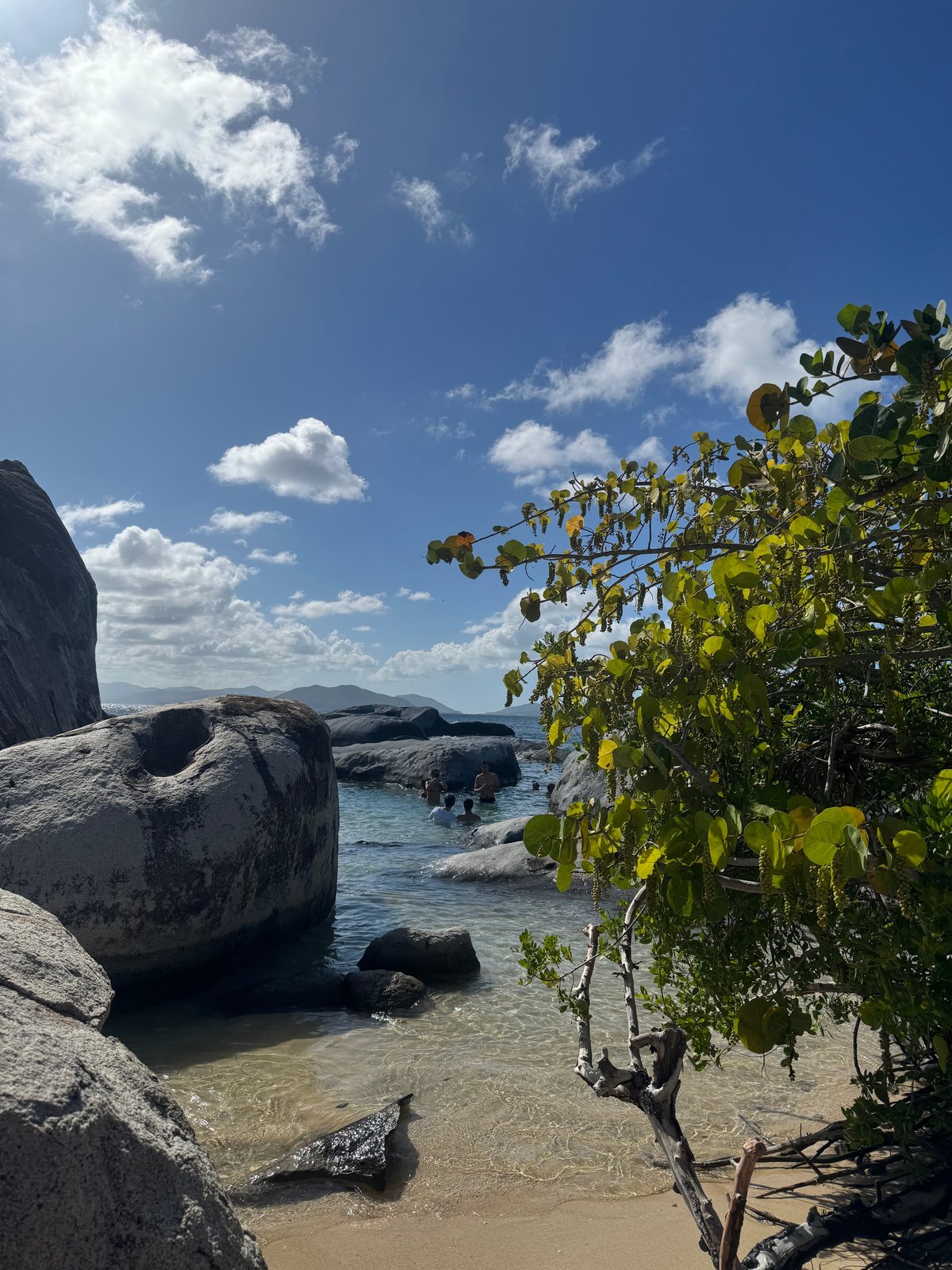 Rocky granite boulders frame a pristine turquoise cove with mangrove vegetation and distant mountains under a bright blue sky.