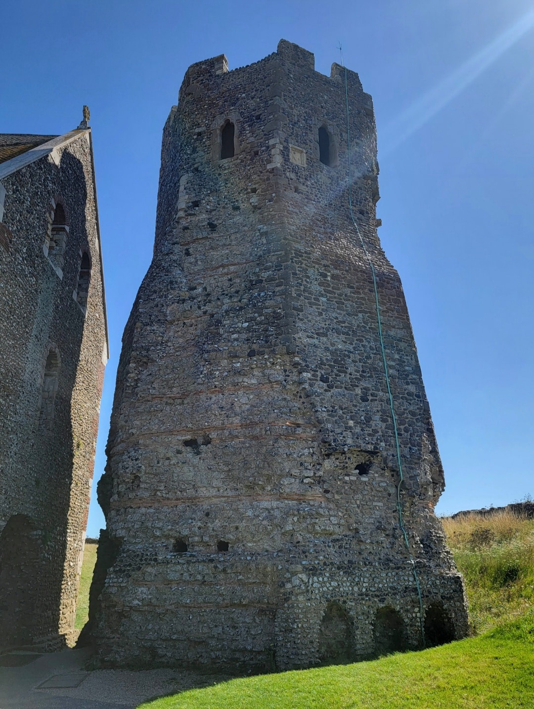 A tall stone round tower with crenellations and narrow windows stands against a blue sky, part of a medieval castle or fortification complex.