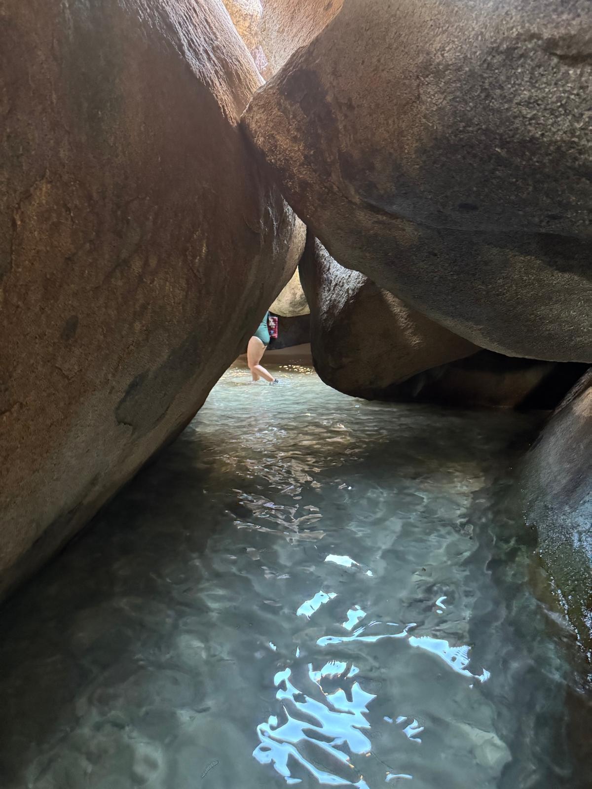 A person wading in clear shallow water between two large granite boulders forming a narrow passage at a tropical beach.
