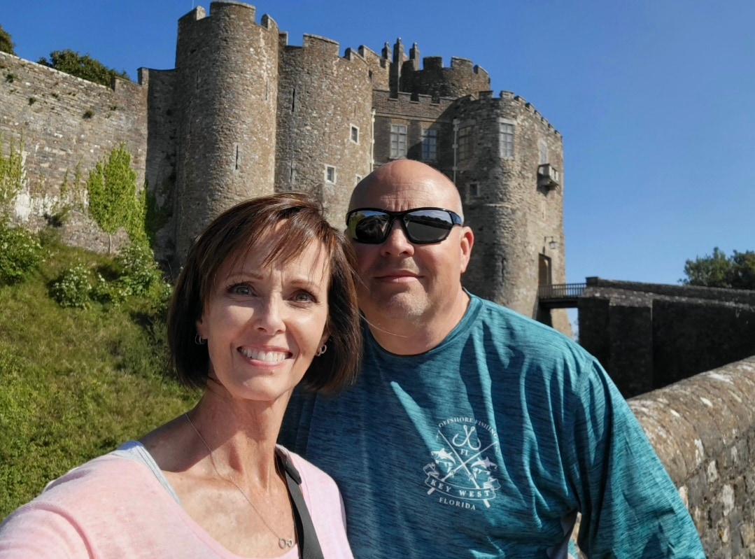 Two adults posing for a photo in front of a stone medieval castle under clear blue skies.