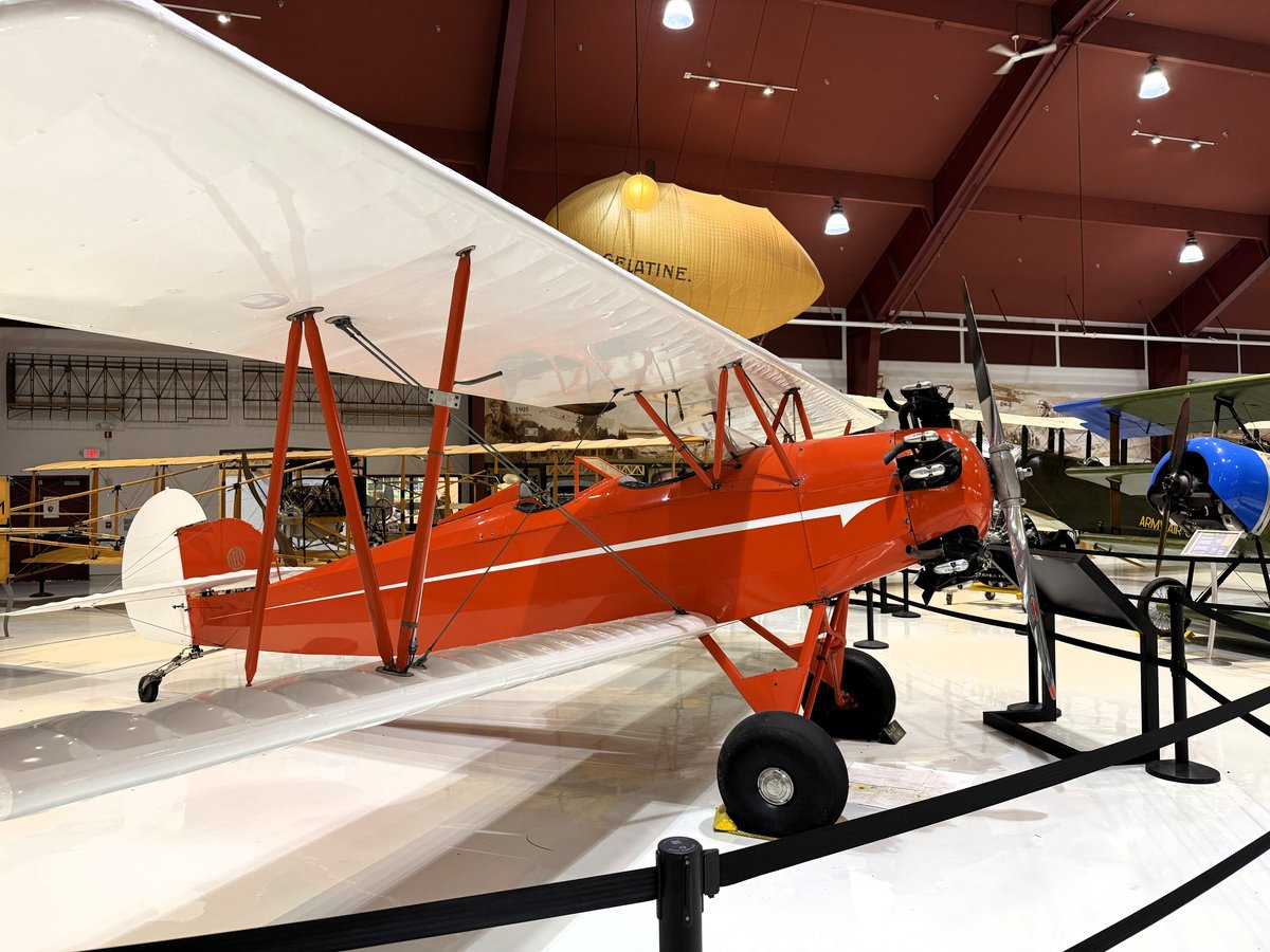 A bright red vintage biplane displayed indoors in a hangar museum with a yellow aircraft visible in the background.