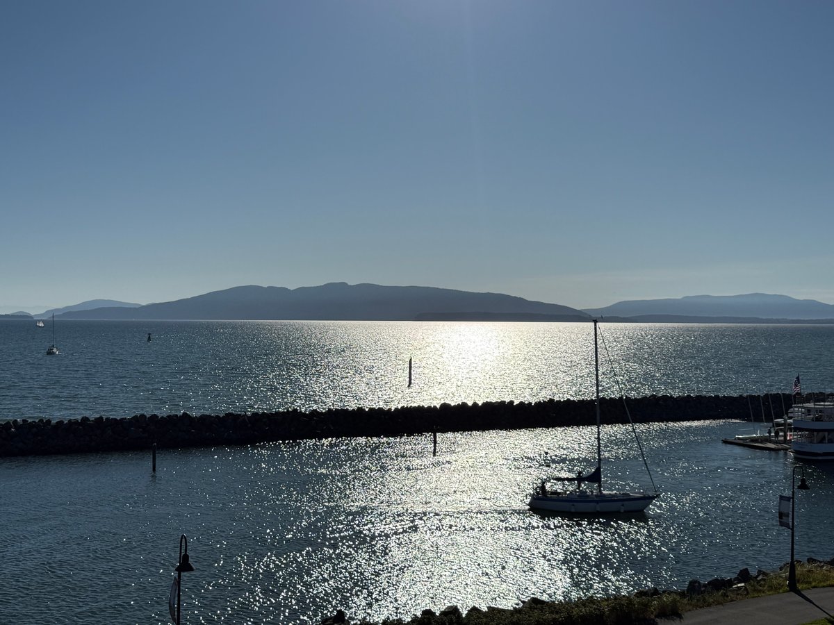 Sailboat moored in a calm harbor with mountains visible across the water under clear blue sky.