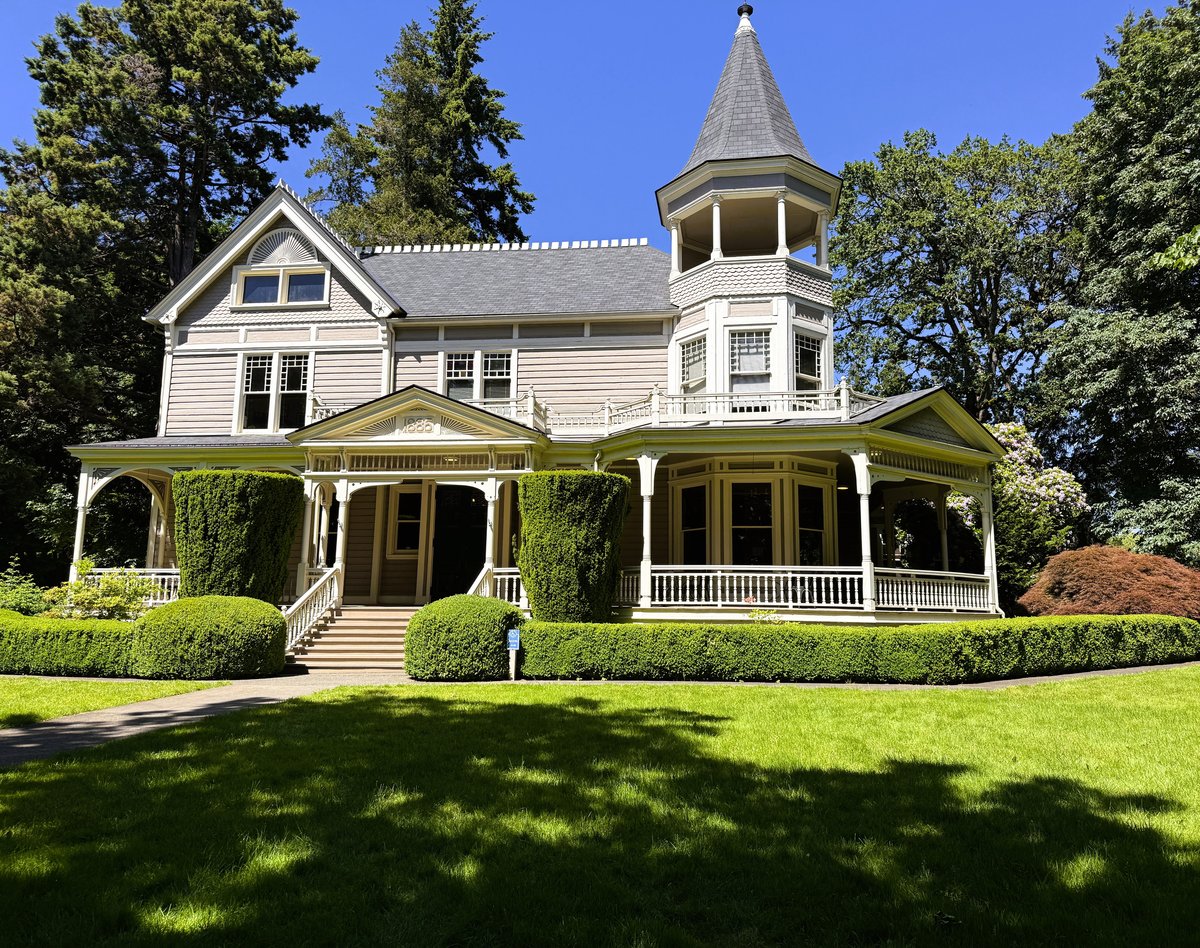 A pristine white Victorian mansion with a distinctive turret and wraparound porch, surrounded by manicured hedges and mature trees on a sunny day.