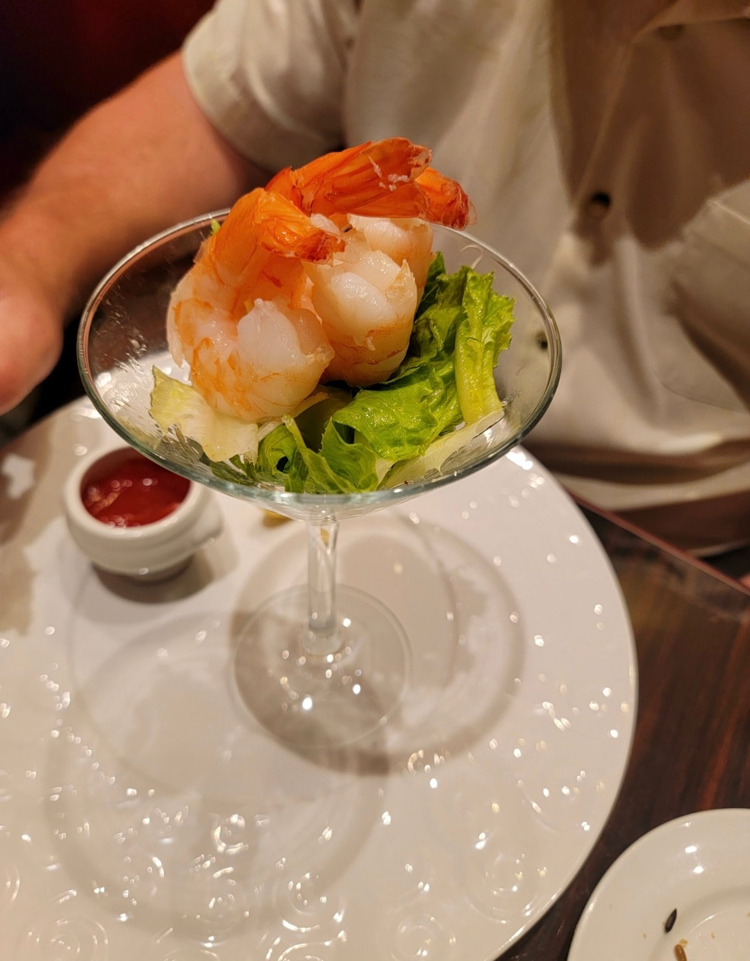 Two large cooked shrimp served in a cocktail glass with fresh greens and a red dipping sauce on a white plate.