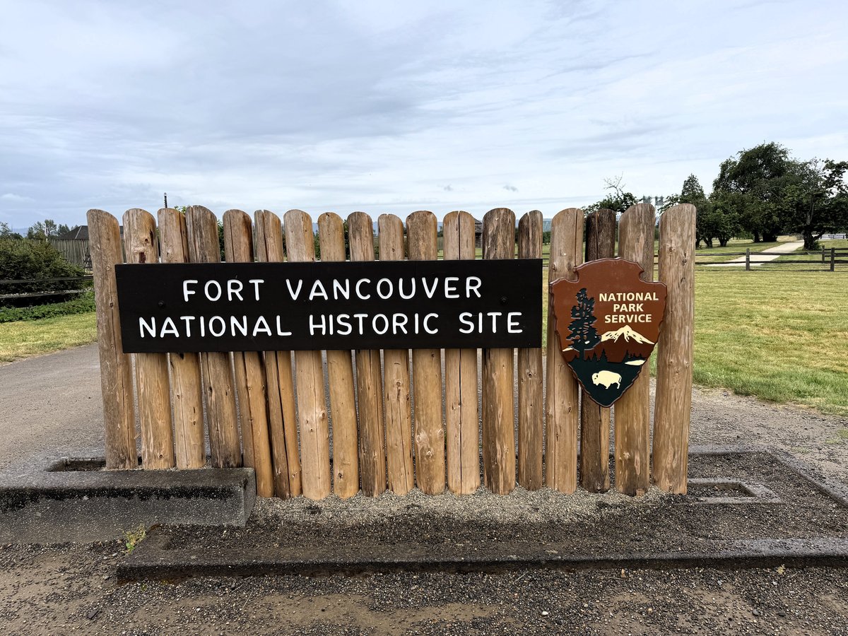 Entrance sign for Fort Vancouver National Historic Site, a wooden palisade structure with National Park Service logo against a grassy field and cloudy sky.