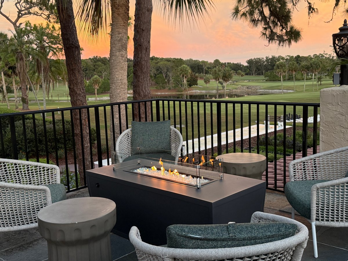 A sunset view from a furnished deck overlooking a golf course and palm-lined landscape with a fire pit table.