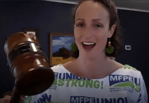 President Amanda Curtis swinging a gavel. She's wearing a blouse decorated with the MFPE logo and colors
