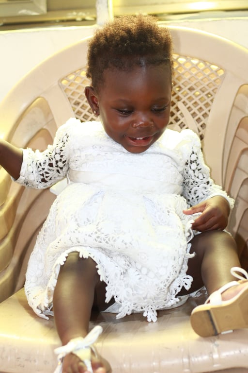 A young child in a white dress seated in a plastic chair