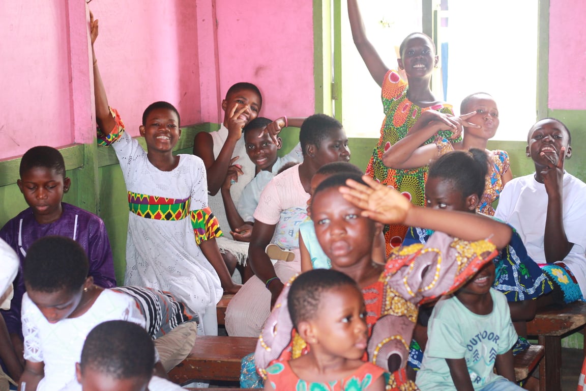 Children gathered in a classroom with pink walls, several raising their hands