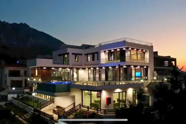 Elegant multi-story luxury mansion illuminated at twilight, nestled near forested mountains.