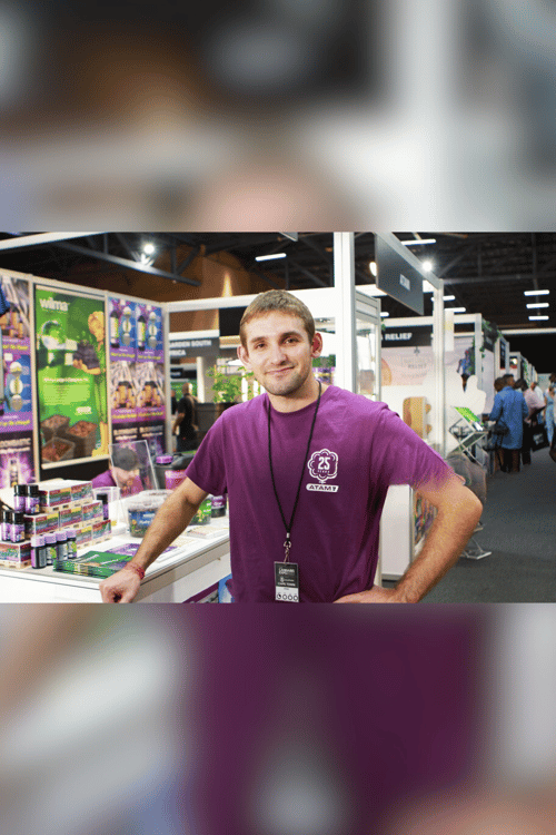 Trade visitor at a cannabis expo – a man in an exhibitor T-shirt surrounded by cultivation products. In the background: exhibition aisles, banners for LED and substrates, a conversational atmosphere. Focus on grow technology, community, expertise.