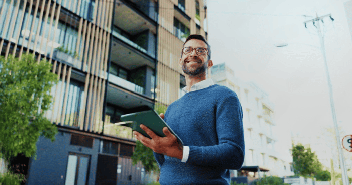 Ein Makler vor einem Mehrfamilienhaus mit Tablet führt Berechnungen für Gebäudeversicherungen bzw. Optimierungen aus.