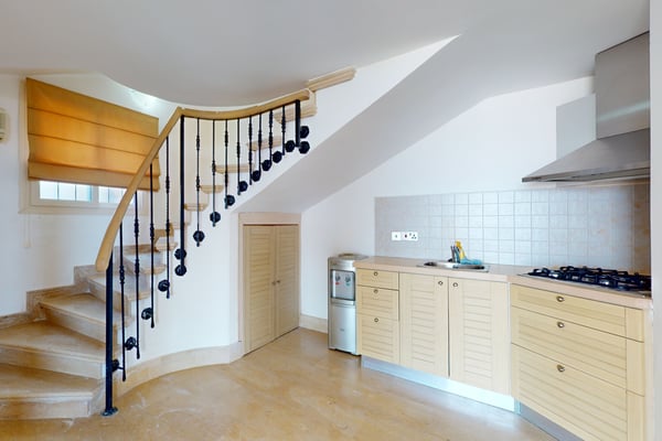 Modern kitchen with wooden cabinets and a gas stove, next to a curved staircase with decorative black railing bars. A small door is located under the stairs, and a water filter stands next to the kitchen counter.