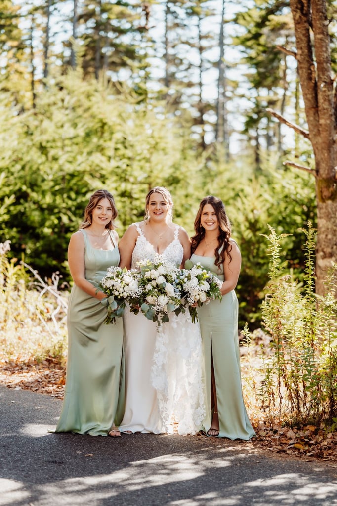 Bride surrounded by two bridesmaids in sage green dresses, all holding white floral bouquets during an outdoor wedding in Acadia National Park, captured by Maine elopement photographer Breezy Photography.