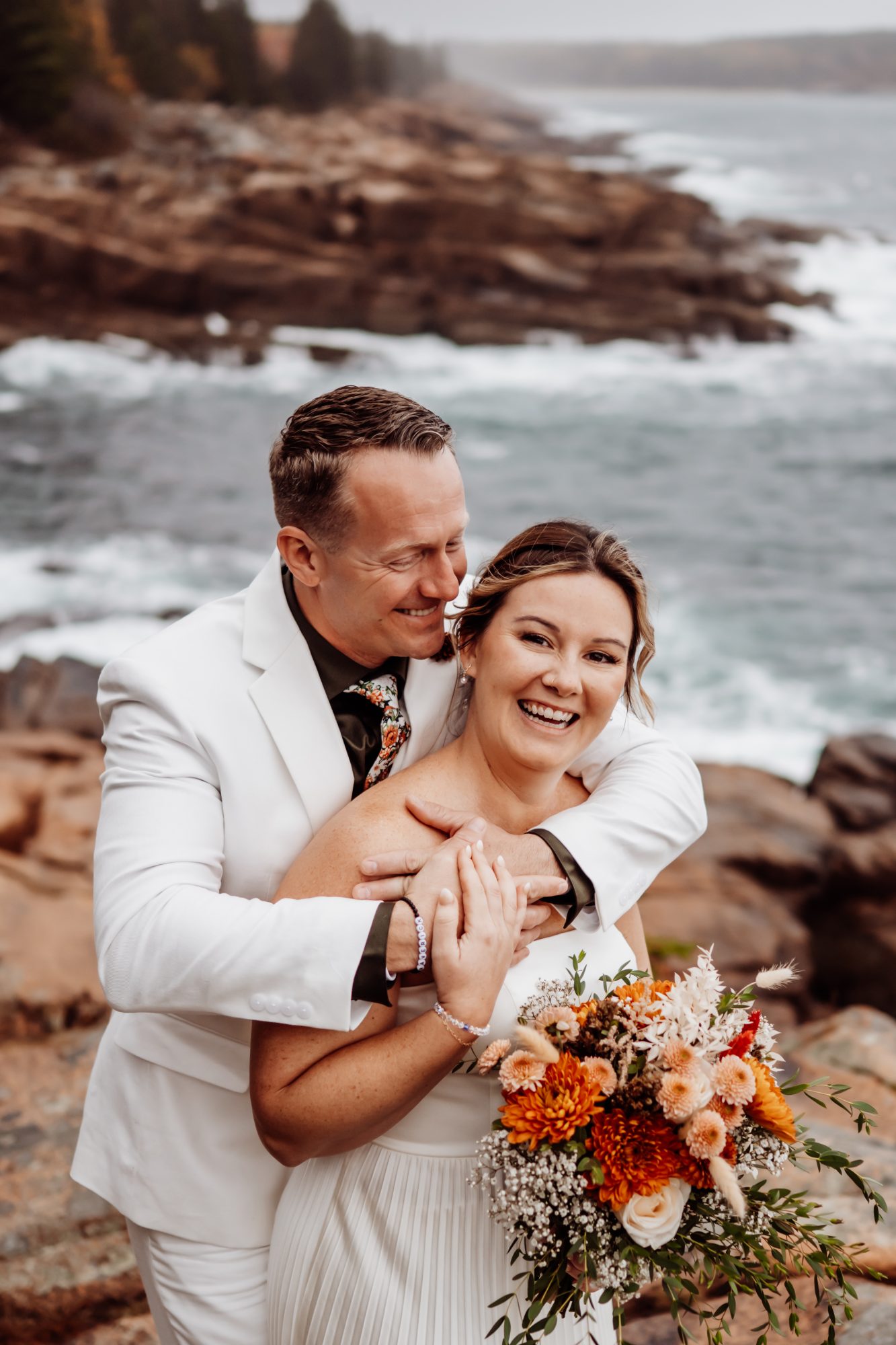Bride and groom laughing together on rocky shoreline at Thunder Hole in Acadia National Park.
