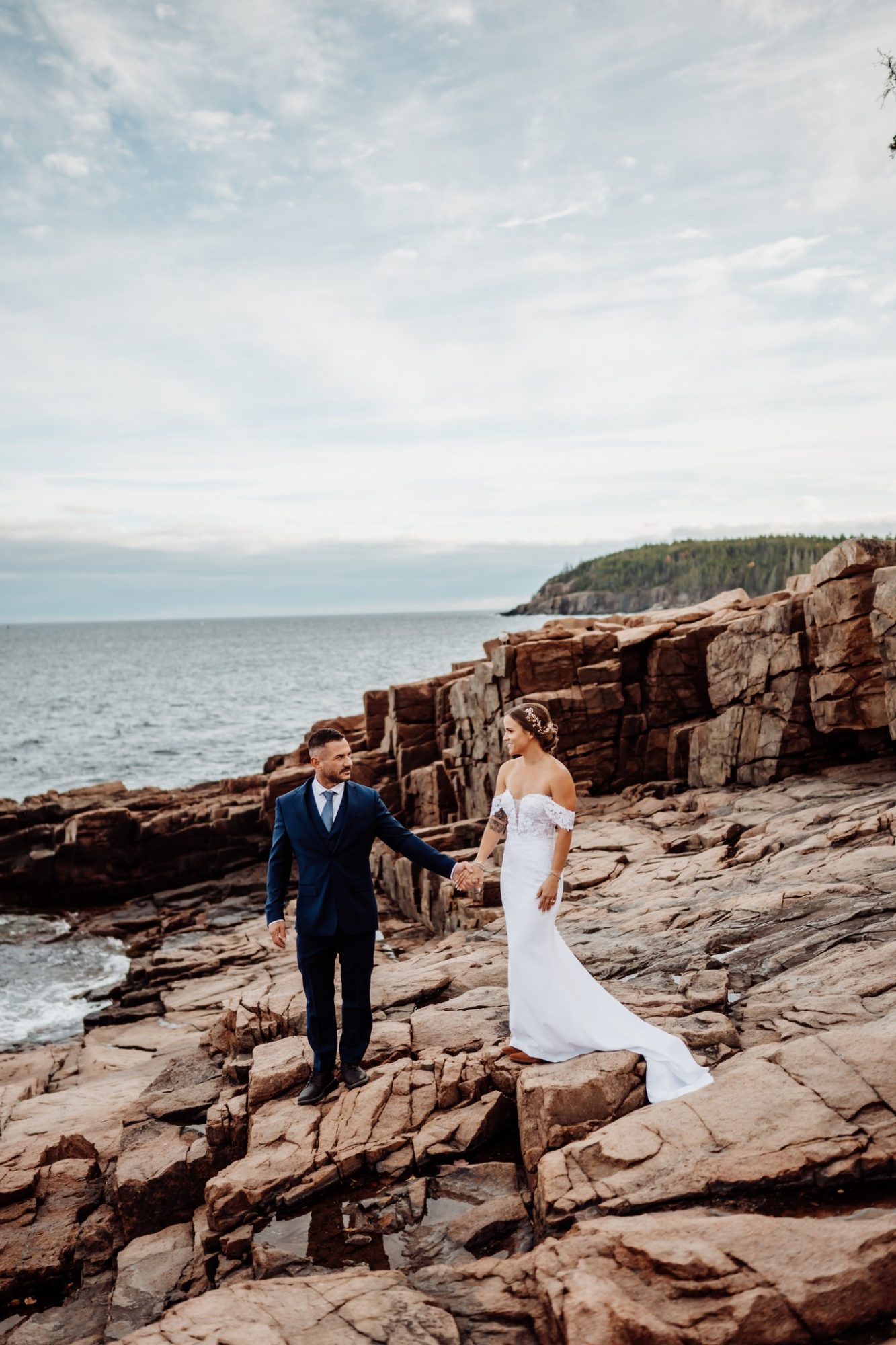 Couple walking hand in hand across the dramatic granite cliffs at Schooner Head Overlook in Acadia National Park, ocean and rugged coastal landscape surrounding their Maine elopement.
