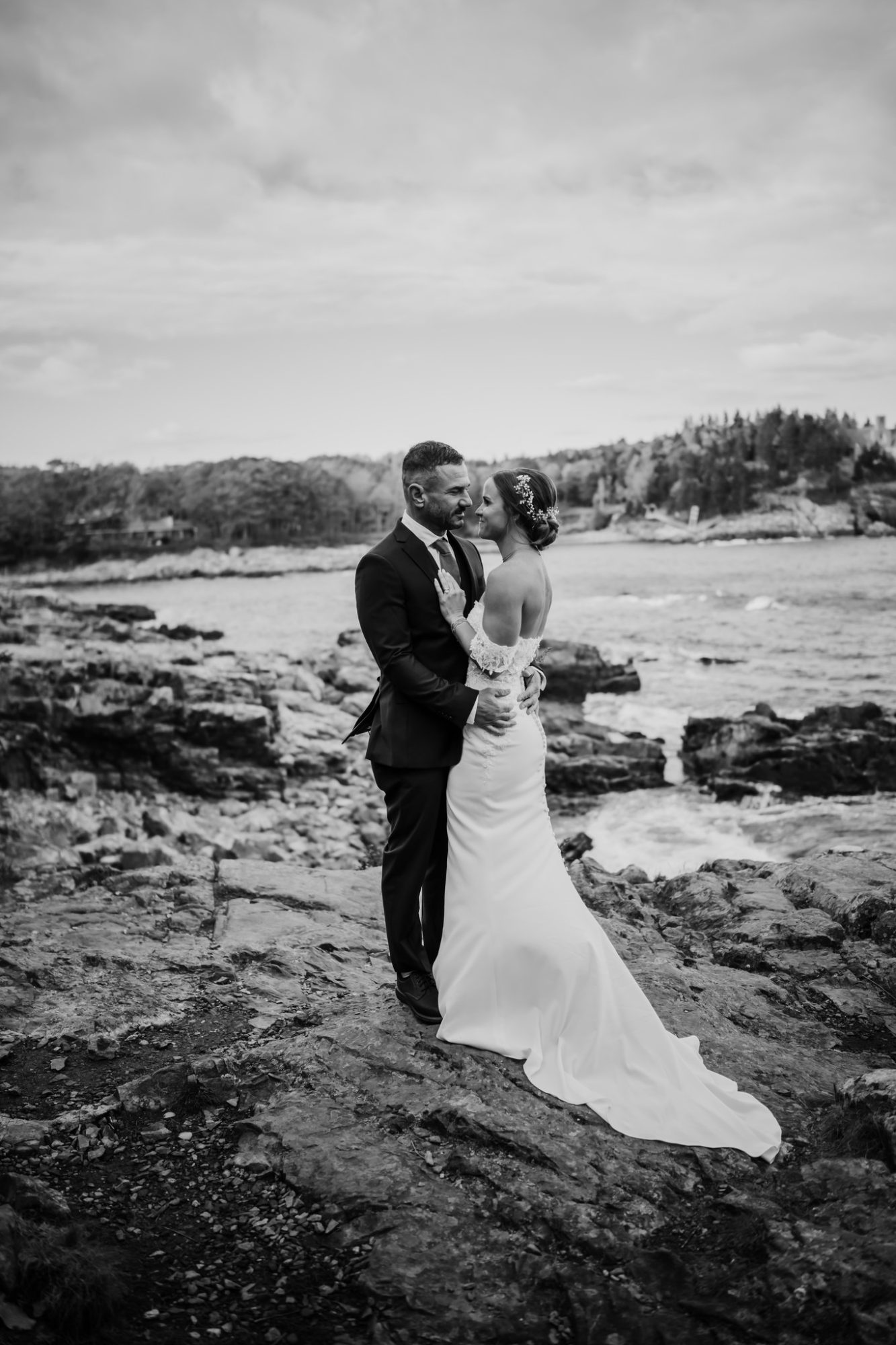 Black and white photo of bride and groom embracing on the rocky shoreline at Schooner Head Overlook in Acadia National Park.