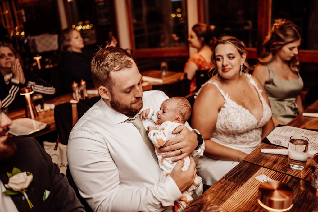 Groom holding a baby while the bride looks on lovingly during their cozy Bar Harbor wedding dinner reception.