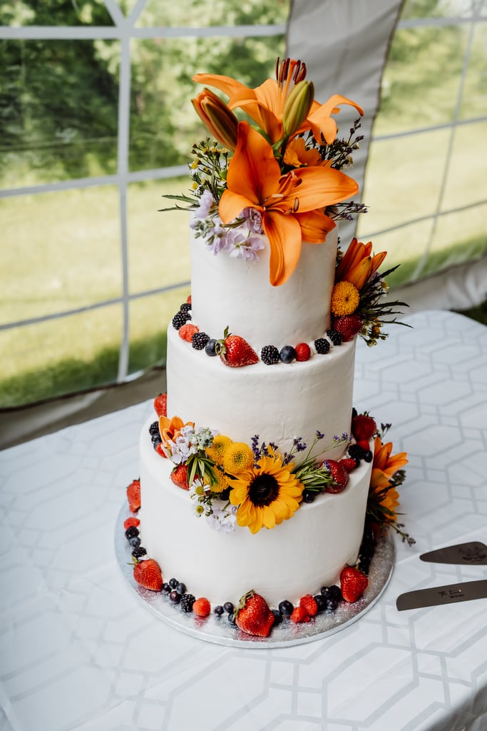 A three-tier wedding cake decorated with vibrant flowers and fresh berries, set on a table under a tent.