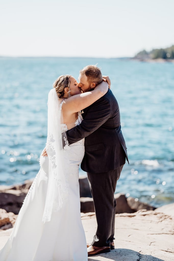 Bride and groom share their first kiss as newlyweds during a seaside wedding at Otter Point in Acadia National Park, sunlight sparkling across the Atlantic Ocean behind them.