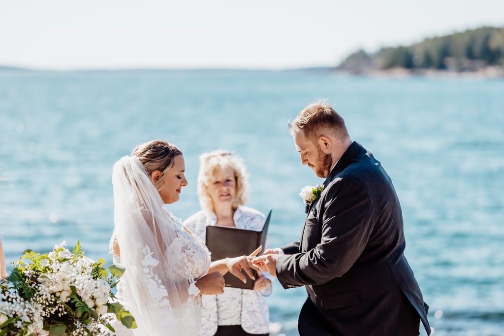 Bride and groom exchanging rings during their oceanfront Acadia National Park ceremony, the officiant smiling as waves glimmer in the background.