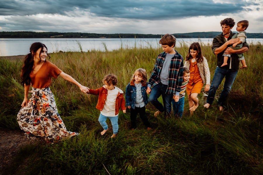 Big family wearing bold colorful outfits walking in a line in grass in front of lake and moody skies