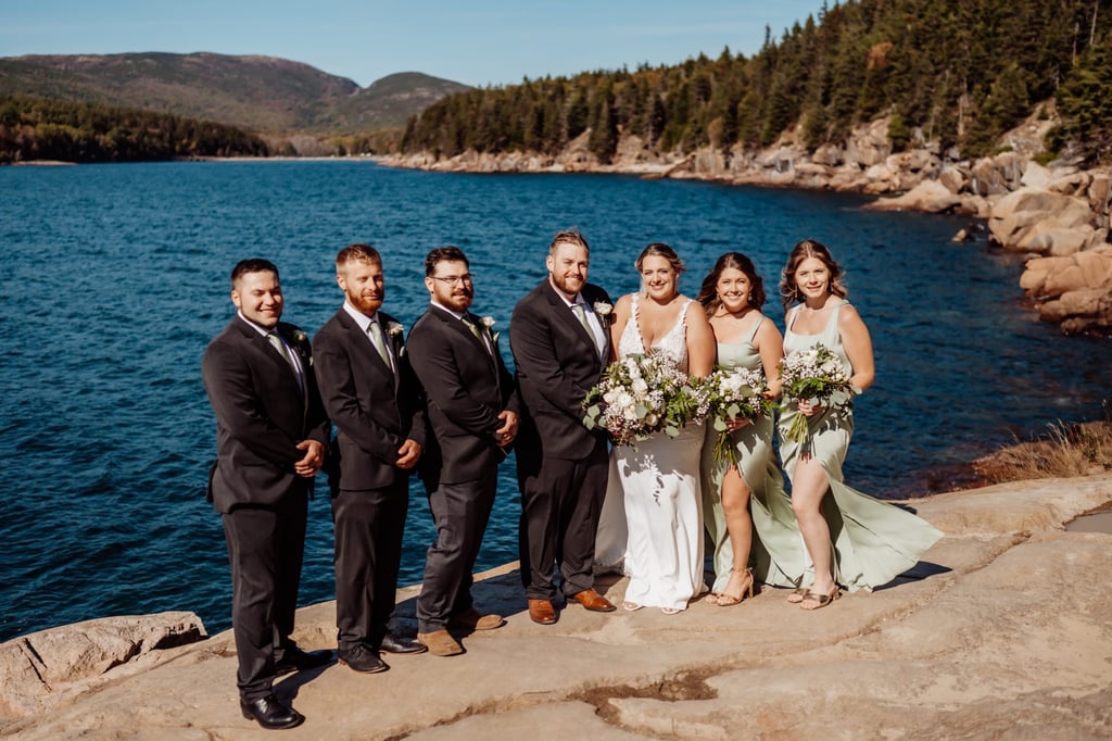 Bridal party smiling together on the rocky shoreline of Acadia National Park, surrounded by pine trees and ocean views during a sunny Maine wedding at Otter Point.