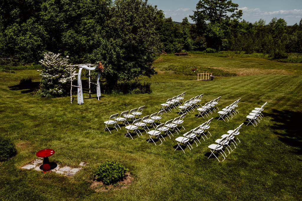 An outdoor wedding ceremony setup with rows of white chairs facing a floral archway, surrounded by lush green trees