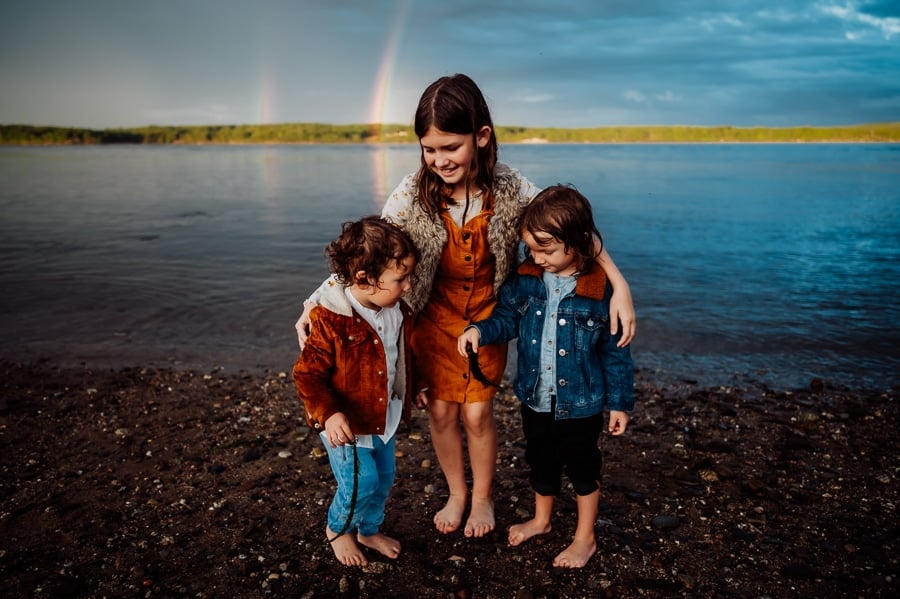 girl in orange dress with two boys in front of double rainbow and lake
