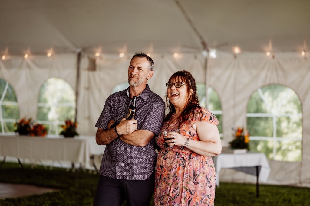 A man and woman enjoying a moment together at a wedding reception, standing under a tent and smiling with drinks in hand.