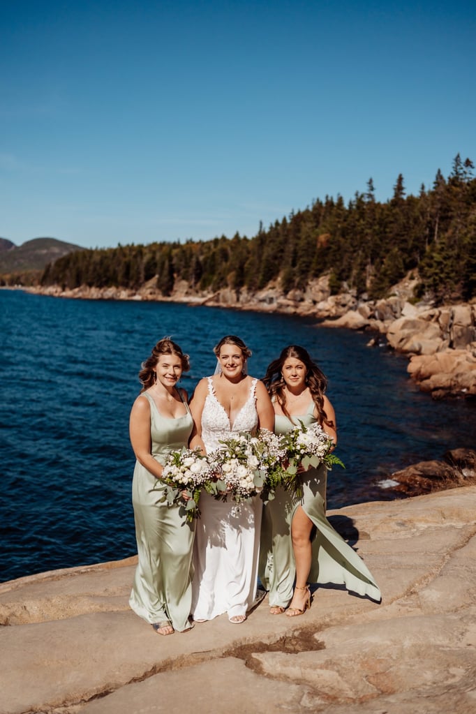 Bride standing with her bridesmaids in sage green dresses, holding white floral bouquets on the rocky cliffs of Otter Point in Acadia National Park with the blue Maine coastline behind them.