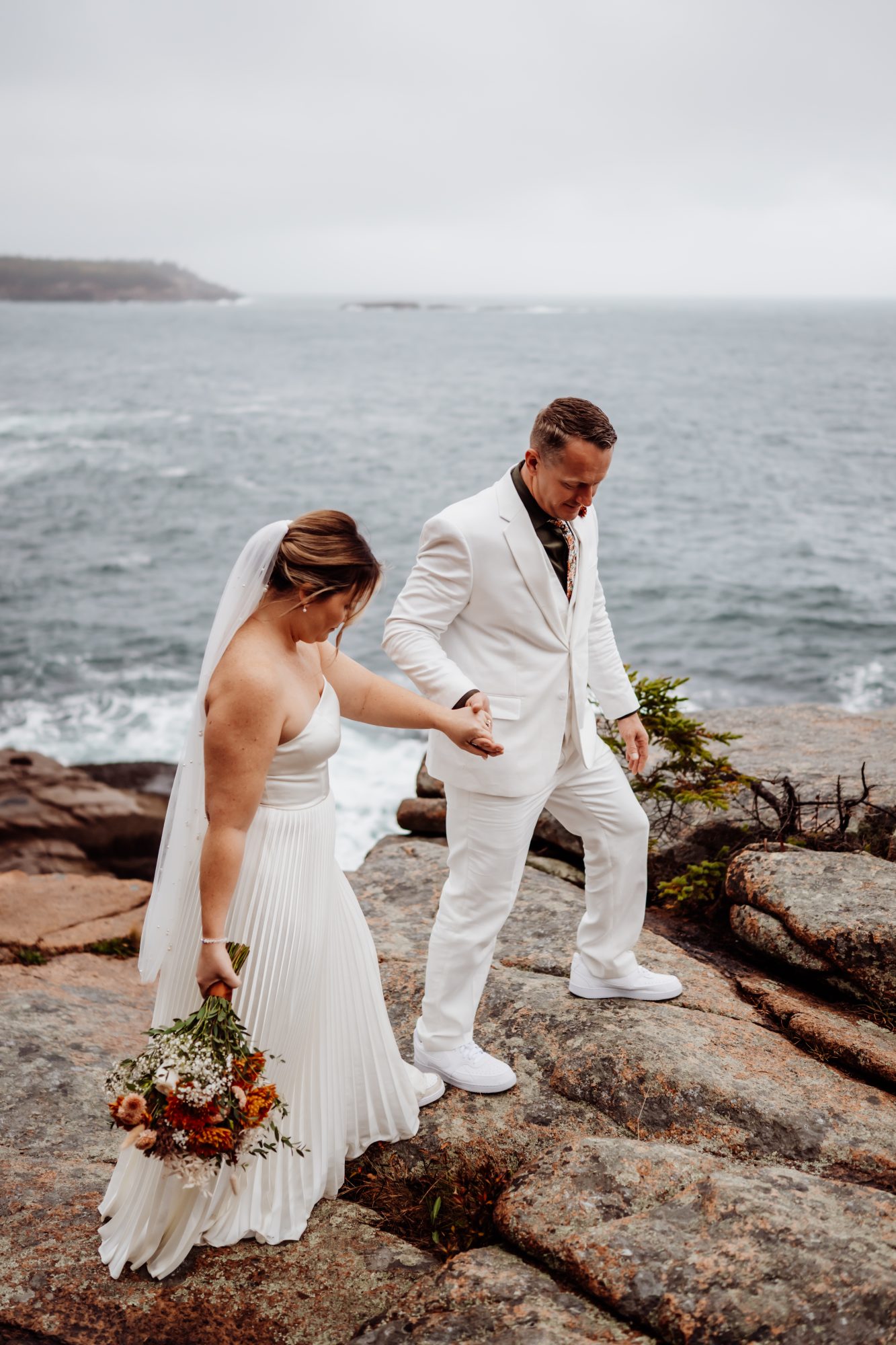 Bride holding bouquet while stepping carefully along rocky ledges at Thunder Hole in Acadia National Park.