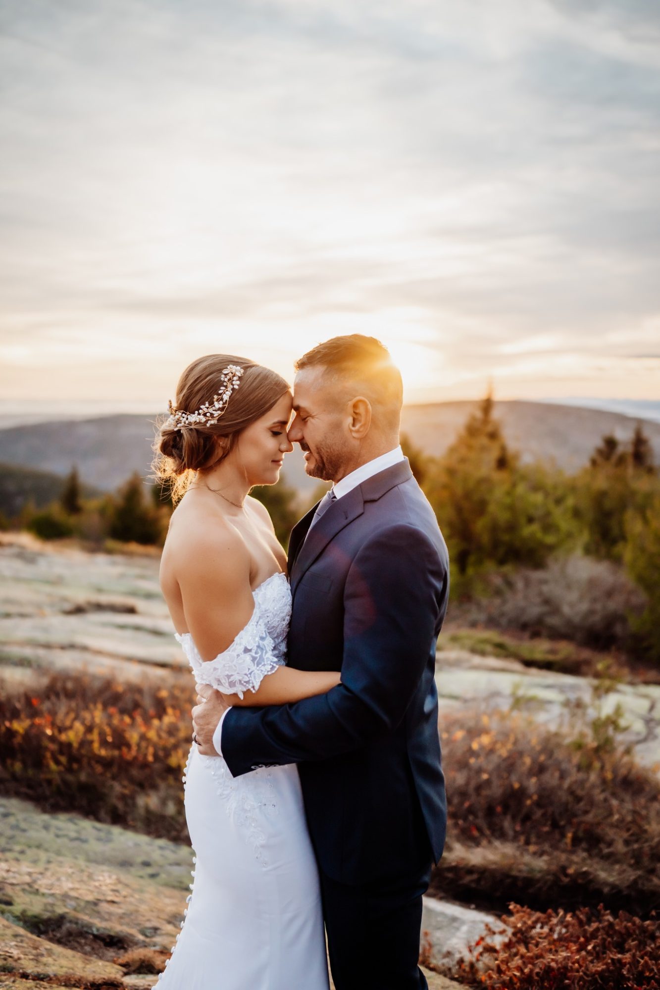 Bride and groom embracing at golden hour after their elopement in Acadia National Park, warm sunset light glowing over the mountains and trees above the Maine coastline.