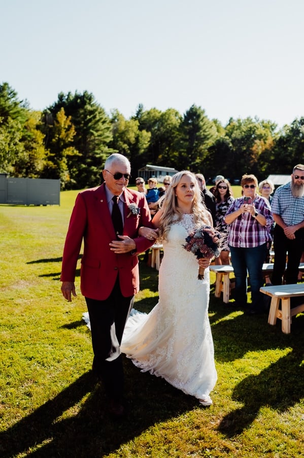 father wearing red tux jacket walking bride down isle at waters edge estate