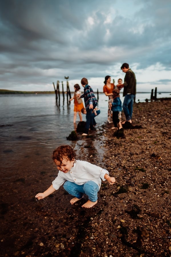 Boy in white shirt bending down and picking up rock on beach with family behind