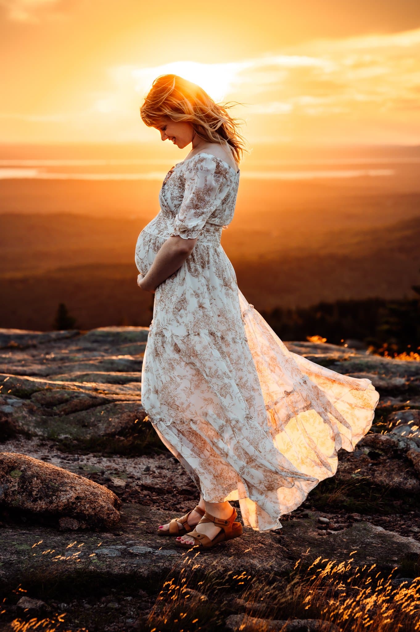 Pregnant woman standing in front of mountain sunset wearing a white dress