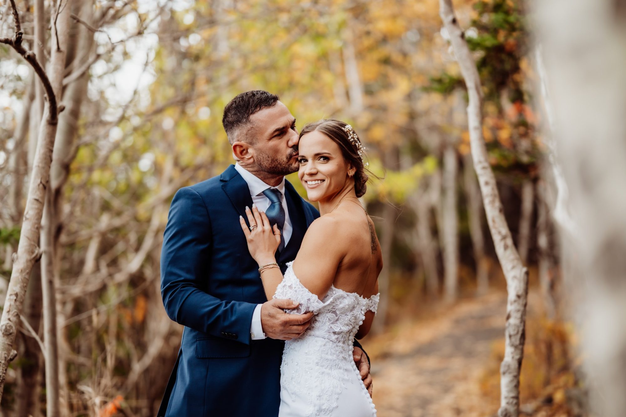 Groom kissing bride’s temple during their fall elopement at Schooner Head Overlook in Acadia National Park surrounded by autumn trees.